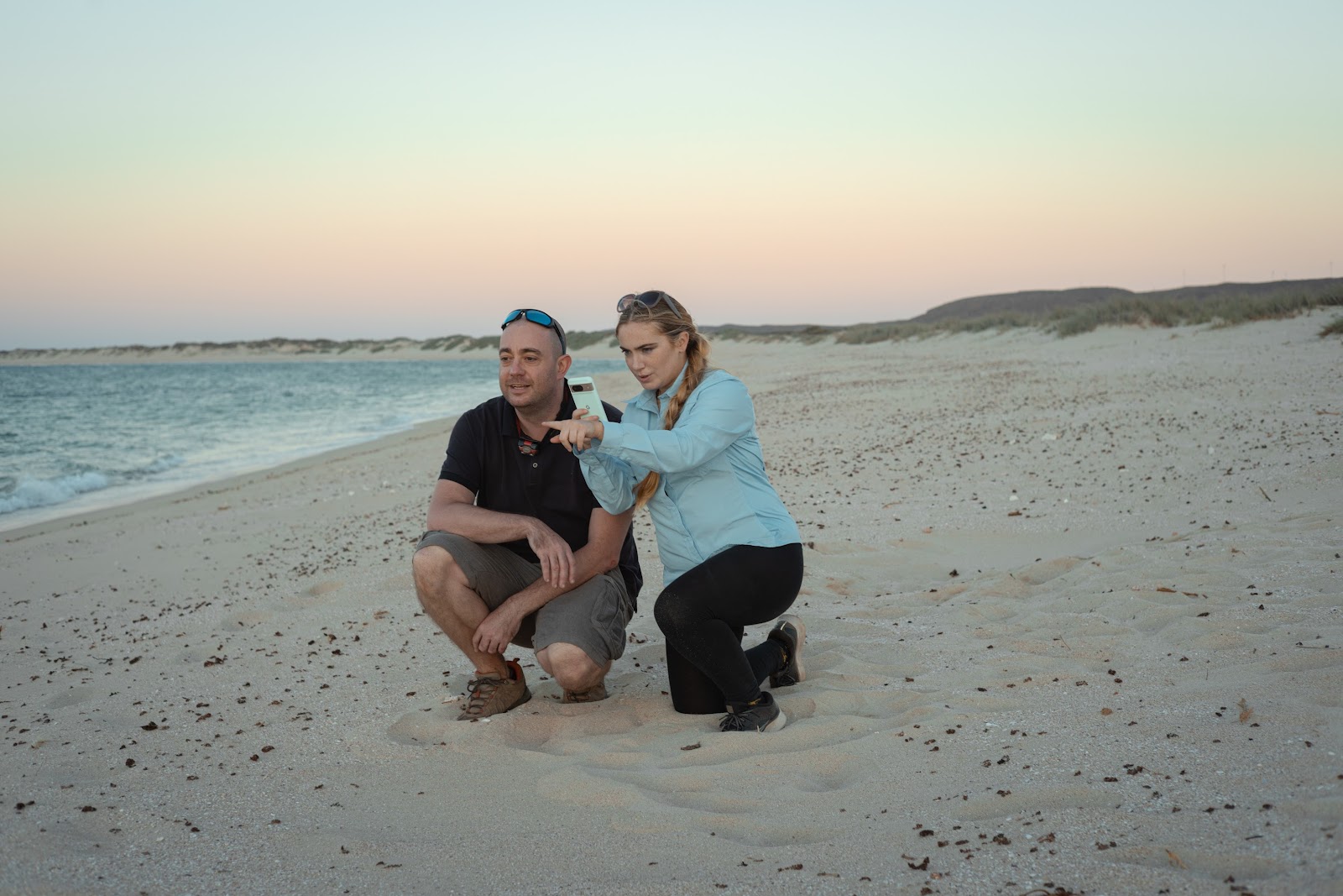 Photo pf Caitlin and Nicolas on the bach in Australia at sunset. Both are kneeling in the sand. Caitlin is using her phone to identify something in the distance, and gesturing to Nicolas who is looking in the same direction