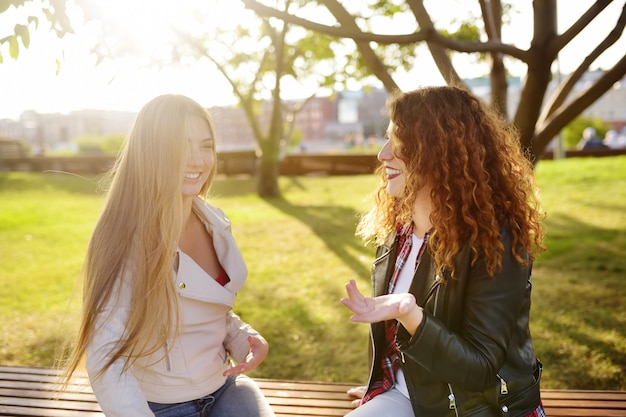 Zwei schöne junge Frauen, die sprechen, während sie auf einer Bank im sonnigen Park sitzen. Kommunikation und Klatsch.