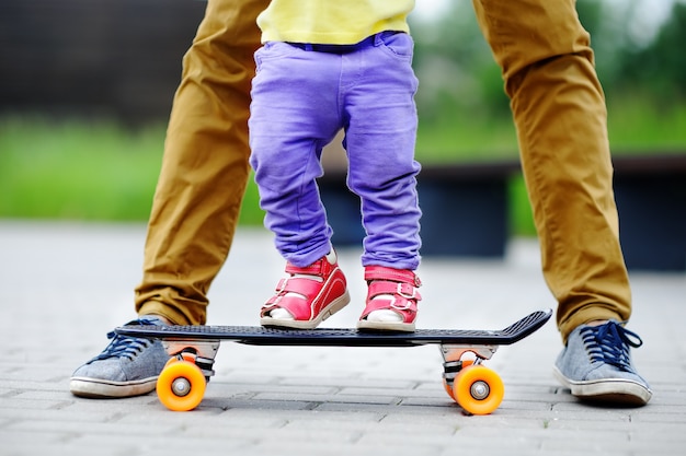 Nettes Kleinkindmädchen, das lernt, mit ihrem Vaterabschluß oben draußen Skateboard zu fahren. Aktiver Familienspaß