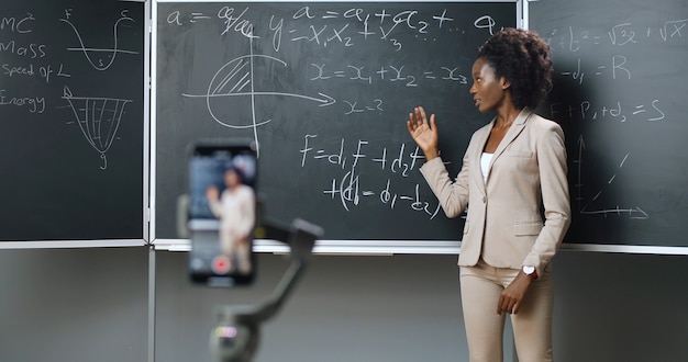 Smartphone recording video lesson at school. Online studying. African American young female teacher explaining math or physics formulas at class. Quarantine concept. Study by internet. Isolation.