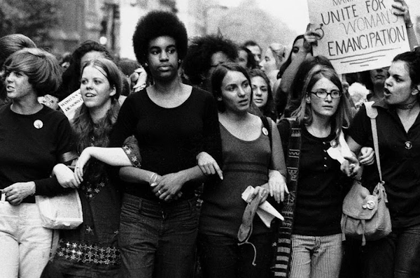 Una multitud de mujeres camina con los brazos entrelazados, cantando y portando pancartas en una manifestación por los derechos de las mujeres.