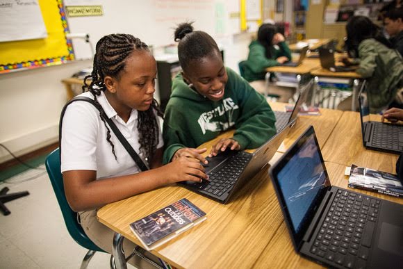 Ortaokul öğrencisi iki kız, sınıfta bir dizüstü bilgisayarı birlikte kullanıyor. Kızlardan polo yaka beyaz tişörtlü olanı ekrana odaklanmış. Yeşil bir kapüşonlu giyen arkadaşı ise ekrana gülümsüyor.
