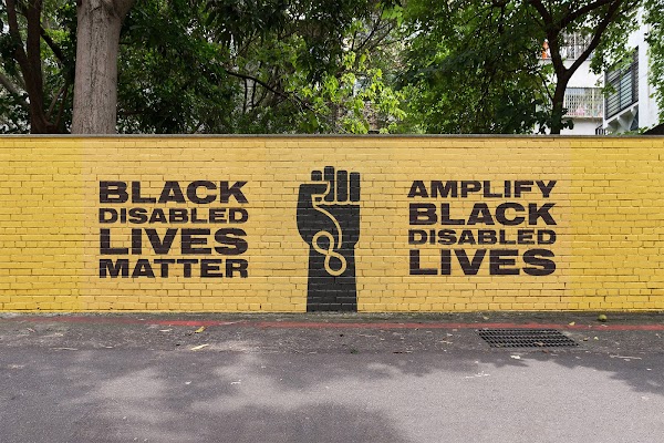 Pared de ladrillo amarilla con las frases "Las vidas de los afrodescendientes discapacitados importan" y "Amplifica las vidas de los afrodescendientes discapacitados" pintadas en negro.