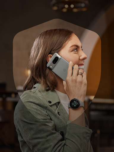 Uśmiechnięta użytkowniczka Androida bezpiecznie odbiera połączenie telefoniczne i widoczny jest jej zegarek Pixel Watch 2. Użytkowniczka otoczona jest ikoną tarczy.