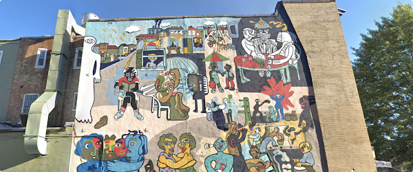 Mural painted on the side of a large building in Washington D.C. with many colors. Subjects in mural are playing games, having meals together, and hugging. Behind the building are large green trees and a blue sky.