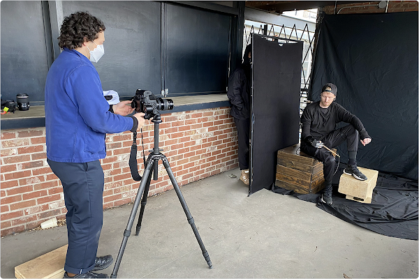 Un hombre con el pelo rizado y una chaqueta azul está detrás de una cámara sobre un trípode. Se trata de Justin Kaneps fotografiando a Jason Barnes, un hombre vestido todo de negro, sobre un fondo negro. Jason es un baterista con discapacidad y sostiene una baqueta accionada por IA.