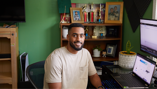 A young man faces the camera and smiles. He is seated in a home office space, with a laptop and monitor