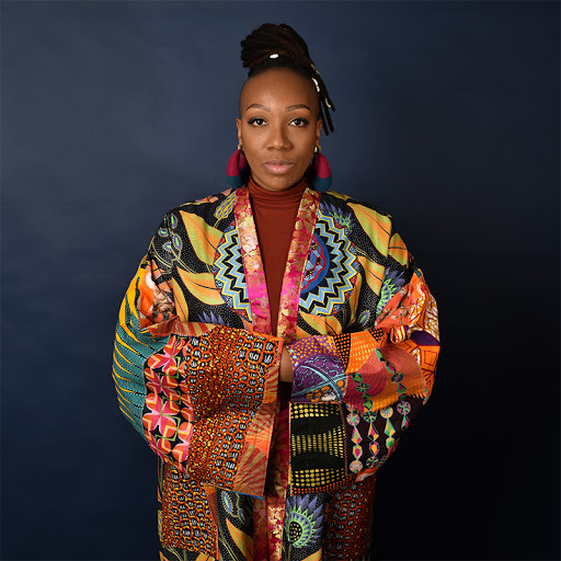 Retrato de la artista con los brazos cruzados vistiendo una chaqueta estilo kimono multicolor con formas geométricas frente a un fondo negro.