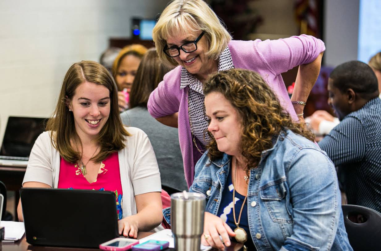 3 glimlachende docenten kijken naar een laptop. Ze bevinden zich in een vergaderruimte waar mensen aan tafels werken op laptops.