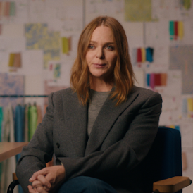 A portrait of fashion designer Stella McCartney in a fashion studio. Several outfits hang on a rack behind her.