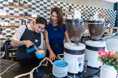 Two women with straight brown hair fill a blue coffee cup behind the counter at a coffee shop.