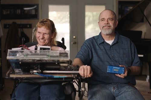 Tania Finlayson sits in a power chair that has a Morse code keyboard. She's smiling and wears a pink shirt with jeans. Her husband, Ken, sits to her left, holding a Morse code device.