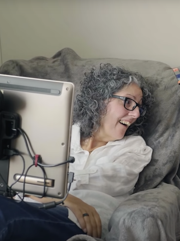 Una mujer con cabello gris y anteojos negros sentada en una silla frente a una pantalla mirando hacia un lado y sonriendo
