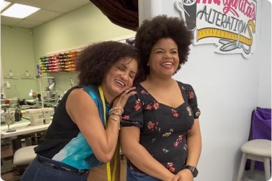 Two women with curly hair embrace at an alterations business, smiling and laughing.