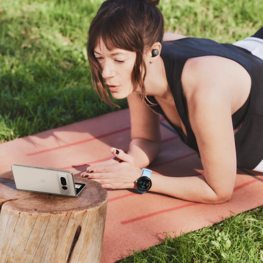 一个人戴着 Wear OS 智能手表和入耳式耳机在瑜伽垫上锻炼，同时看着一部 Android 可折叠手机。