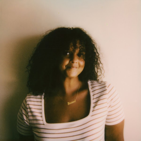 Retrato de la artista en un top de cuello redondo a rayas marrón y crema delante de un fondo beige.