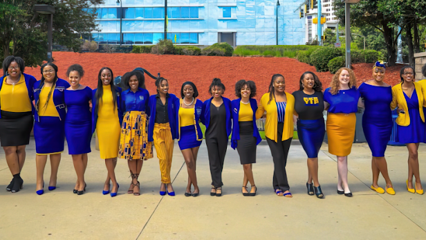 Un grupo de 16 mujeres afrodescendientes vestidas de azul y amarillo están de pie en una fila y sonríen a la cámara.