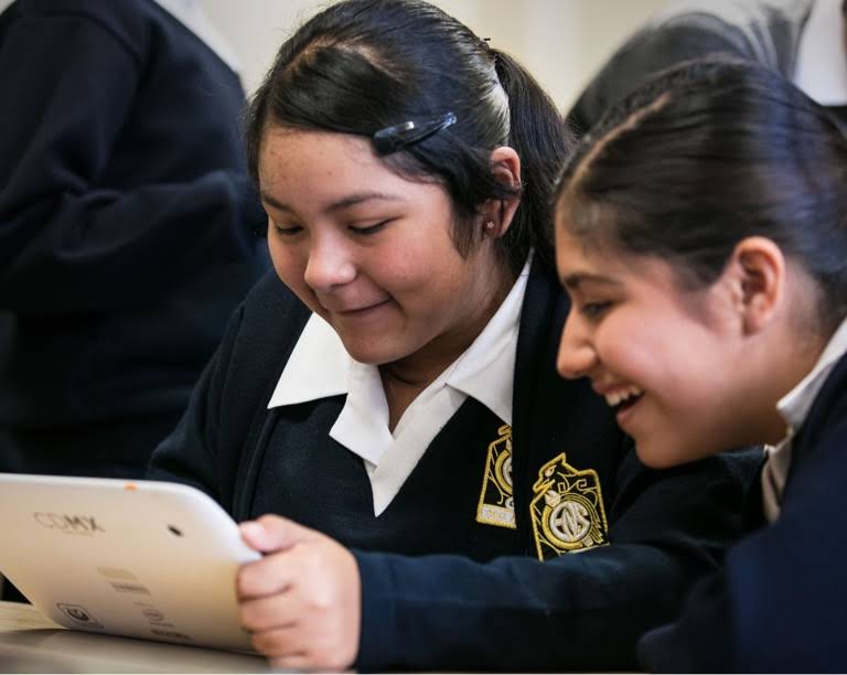 To piger i skoleuniform smiler, og en af dem har en tablet i hånden