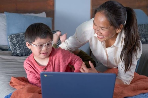 Un niño con anteojos usa una computadora portátil sobre una cama con su mamá