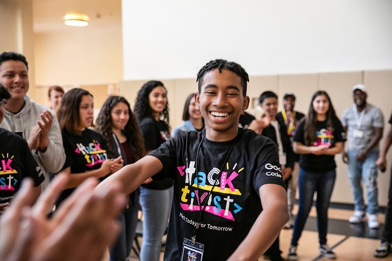 Teenager in black Hacktivist shirt smiling, surrounded by group of teens in school gymnasium.