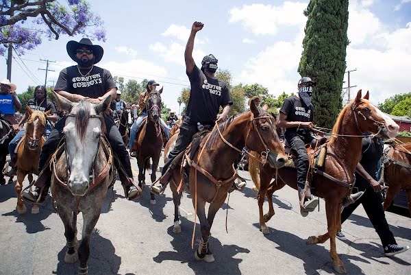 Randy and The Compton Cowboys leading a group of thousands on horseback through the streets of Compton wearing Compton Cowboy merch, cowboy hats, and fists raised.