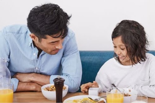 朝食の席で父親が子どもの横に座り、白いデバイスを使っている子どもを見ています。子どもは笑顔で白いデバイスを使っています。