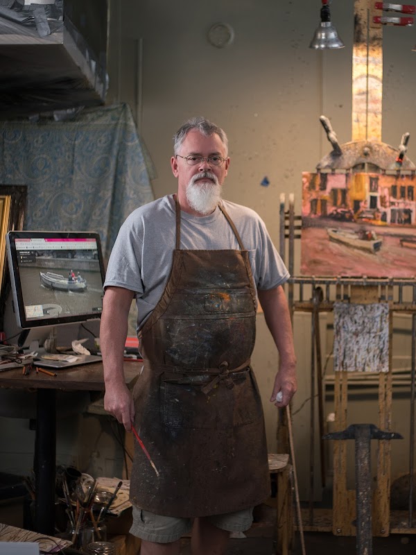 Painter Bill Guffey stands in his art studio. He has a white beard and is wearing a brown apron with paint stains.