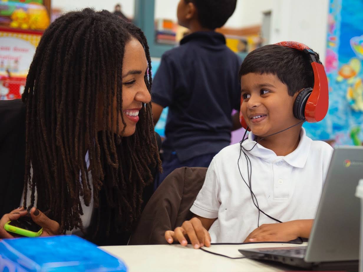 En kvinna lutar sig fram och ler mot en elev som använder en Chromebook med hörlurar.