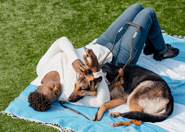 Une femme noire est allongée sur une couverture dans l'herbe avec son chien d'assistance, un berger allemand. Elle tient son téléphone Android à la main.