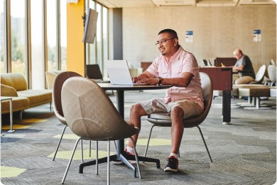 A man in glasses, a pink button down shirt and shorts sits at his computer a table in a workspace.