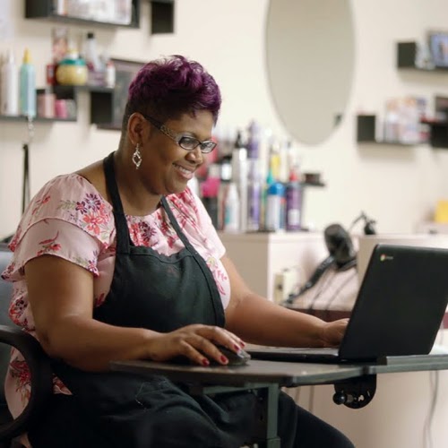 A hairdresser smiles while she uses a laptop on a small table in the salon.