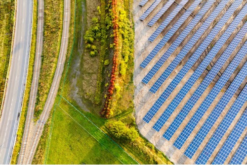 A bird’s eye view of a field covered in solar panels. To the left edge of the lot, there’s greenery with trees.