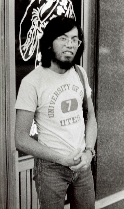 Black and white photo of Corky with long black hair, wearing a t shirt, and holding a camera over his shoulder