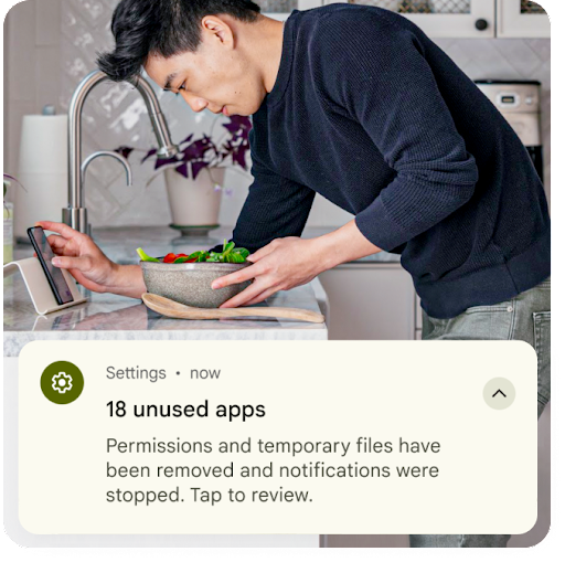 En person laver mad ved køkkenvasken og kigger samtidigt på sin Android-telefon. En grafisk overlejring af en notifikation fra Indstillinger er placeret hen over billedet. Der står, at midlertidige filer fra apps, der ikke bruges, er blevet fjernet, og at tilladelserne er nulstillet.