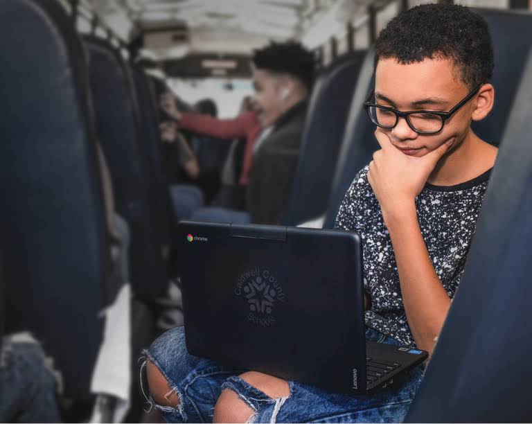A student wearing glasses, seated, focused on a Chromebook device on a bus while commuting